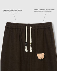 Close-up of elastic waistband with cream drawstring cords and plush teddy bear face on dark brown wool drape pants with fabric callouts
