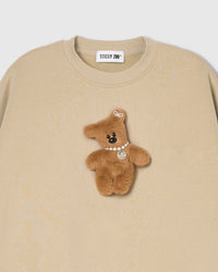 Close-up of plush teddy bear with pearl necklace and ear bow on cream cotton sweatshirt showing 3D texture
