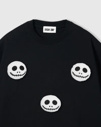 Close-up of plush skull face showing white plush body, hollow eyes, and stitched grin on black cotton