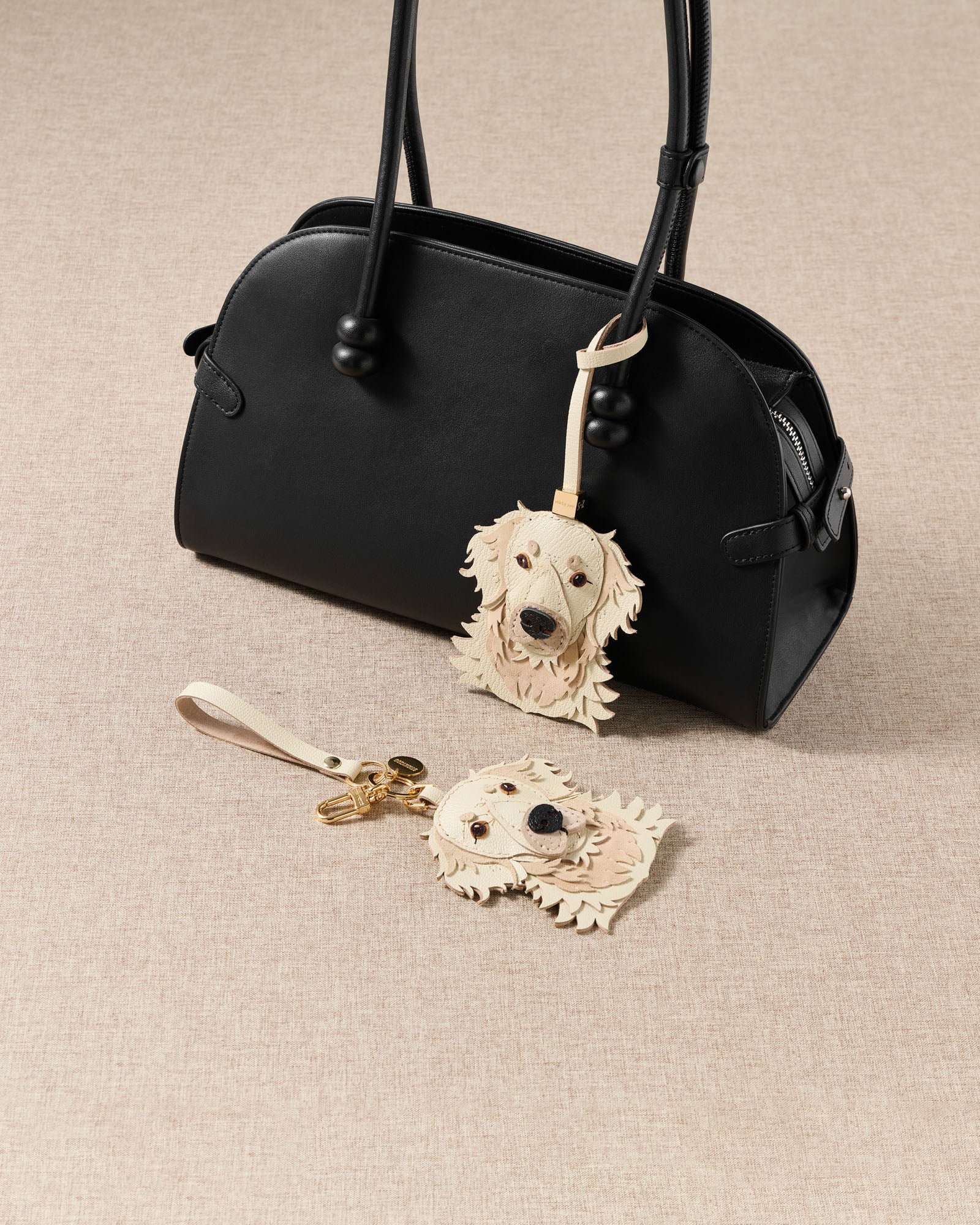 {{Strap loop or keychain clip.}} Both cream Golden Retriever charm editions with black bag — handmade French Alran goatskin with 18K gold-plated hardware. 72 hours of craft per piece.