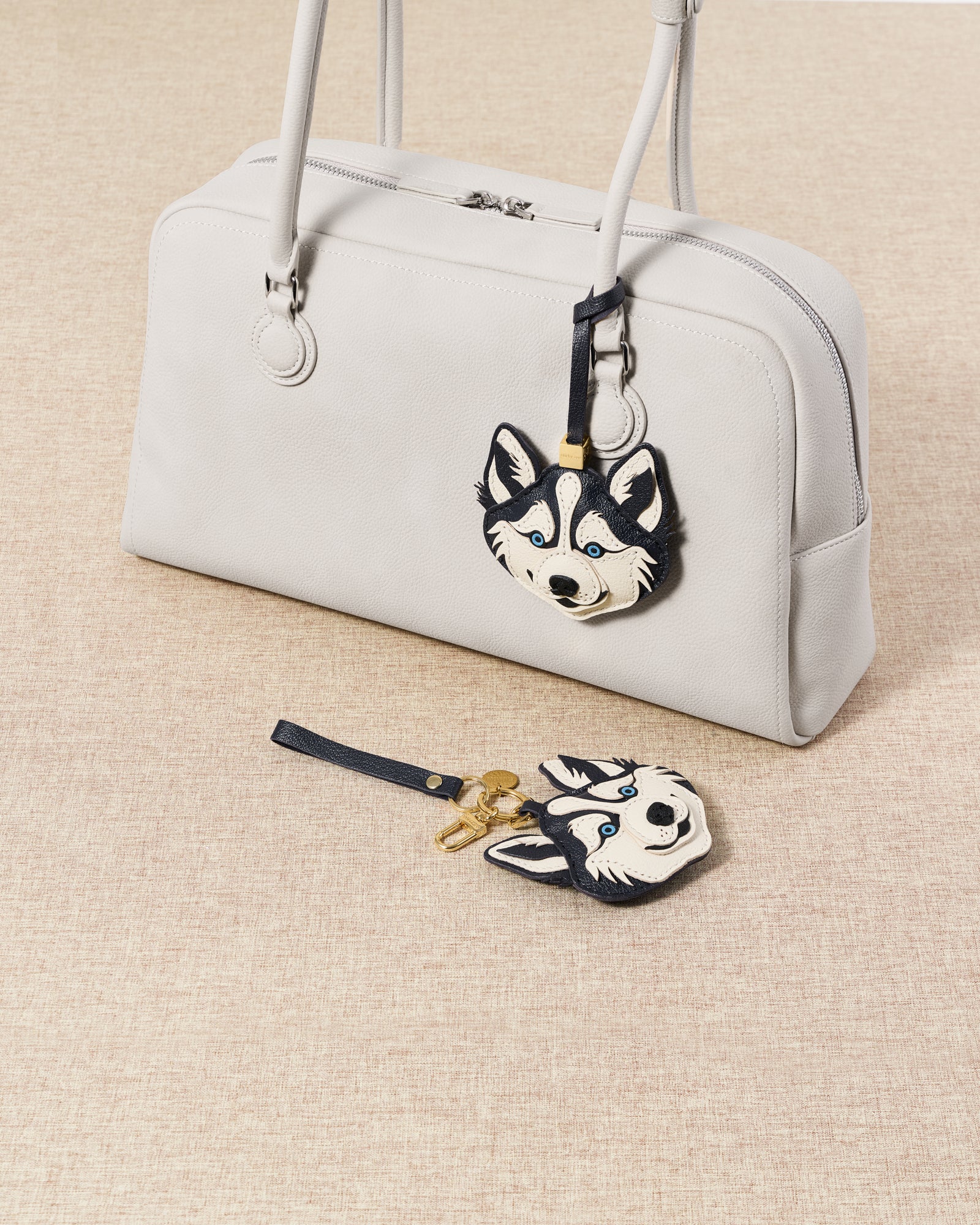 {{Designed in Amsterdam}} Two husky charms with grey bag and blue bag flat-lay on beige linen surface