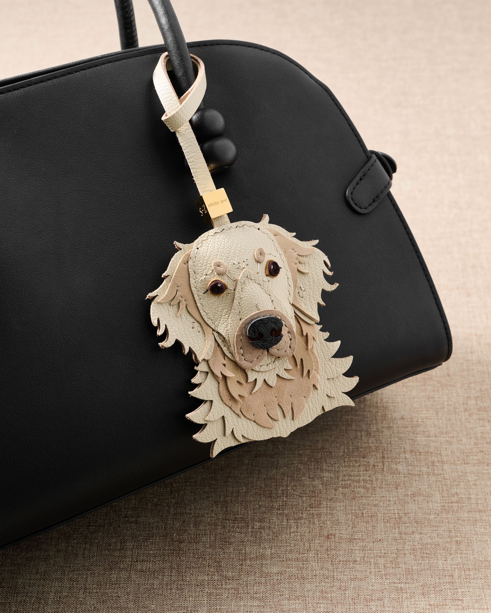 {{Designed in Amsterdam}} Styled view of cream golden retriever charm showing full leather strap and engraved gold STICKYZOO hardware