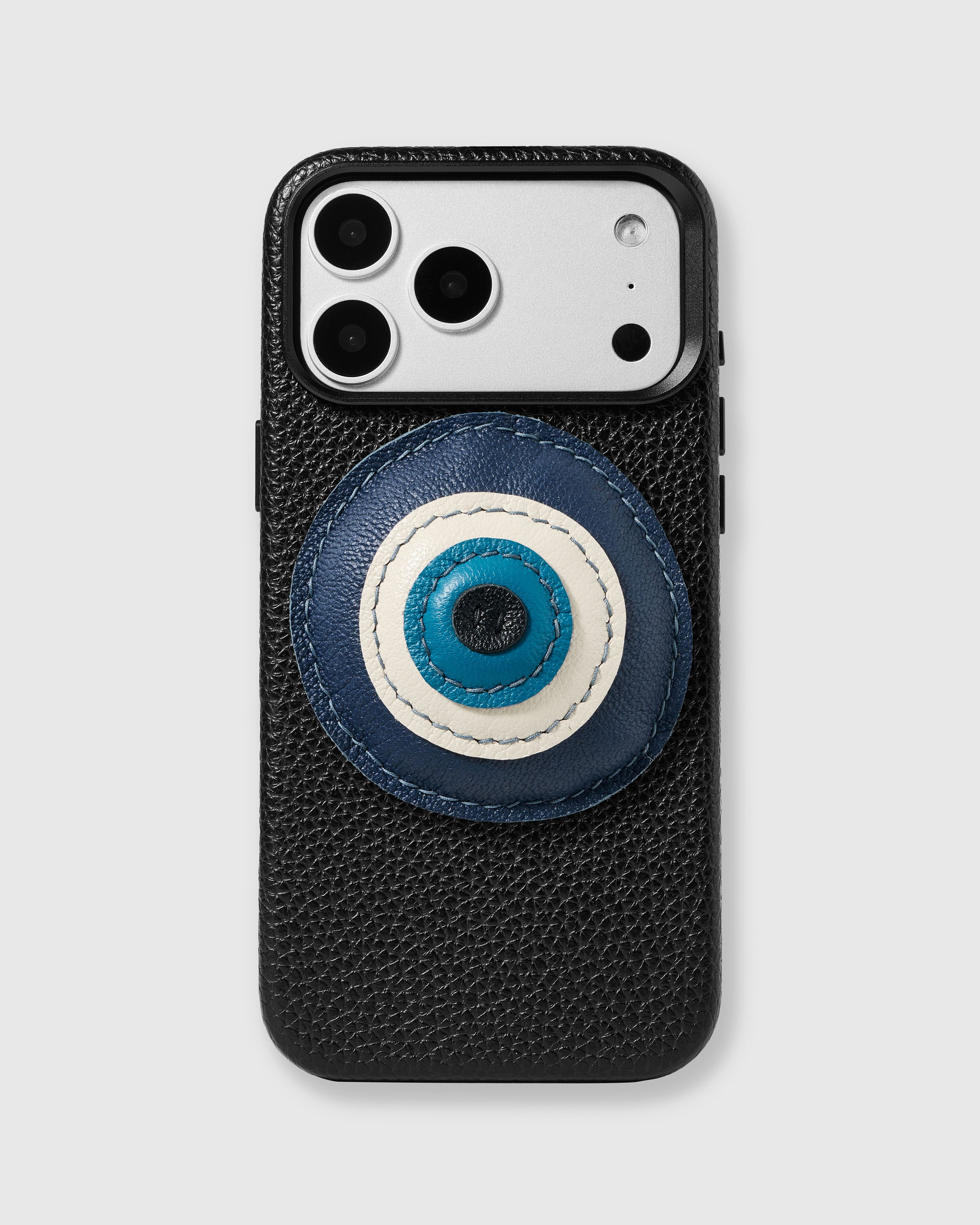 {{Hand-stitched leather evil eye.}} STICKYZOO Evil Eye Charm Case in black with 3D handmade French Alran leather evil eye charm in layered navy, ivory, turquoise, and black concentric circles.