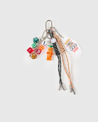 STICKYZOO Lucky Harvest Stack Charm flat lay with carrot-suited collectible figure, colorful polyhedral dice, Always Bee Kind tag, and dark green and peach hand-braided paracord ropes with stainless steel metal tips