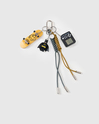 Handmade stack charm | STICKYZOO Shadow Stack Charm flat lay featuring yellow fingerboard skateboard deck, dark caped hero collectible figure, retro mini game player console, and black and mustard hand-braided paracord ropes with stainless steel metal tips