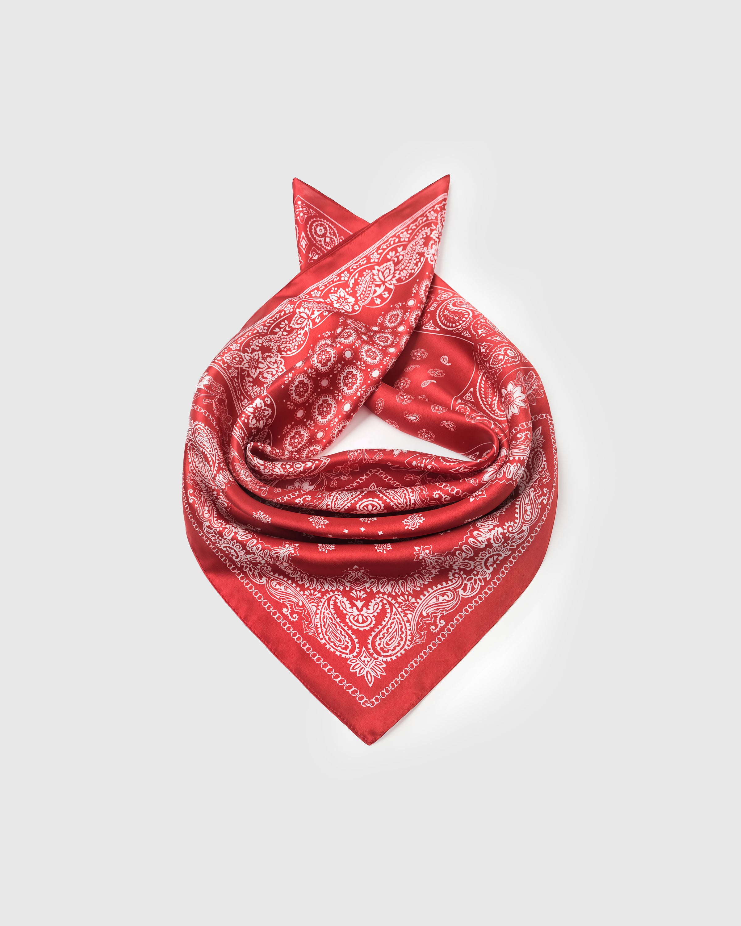Red bandana silk scarf coiled on a white surface showing the ivory line-work and silk drape