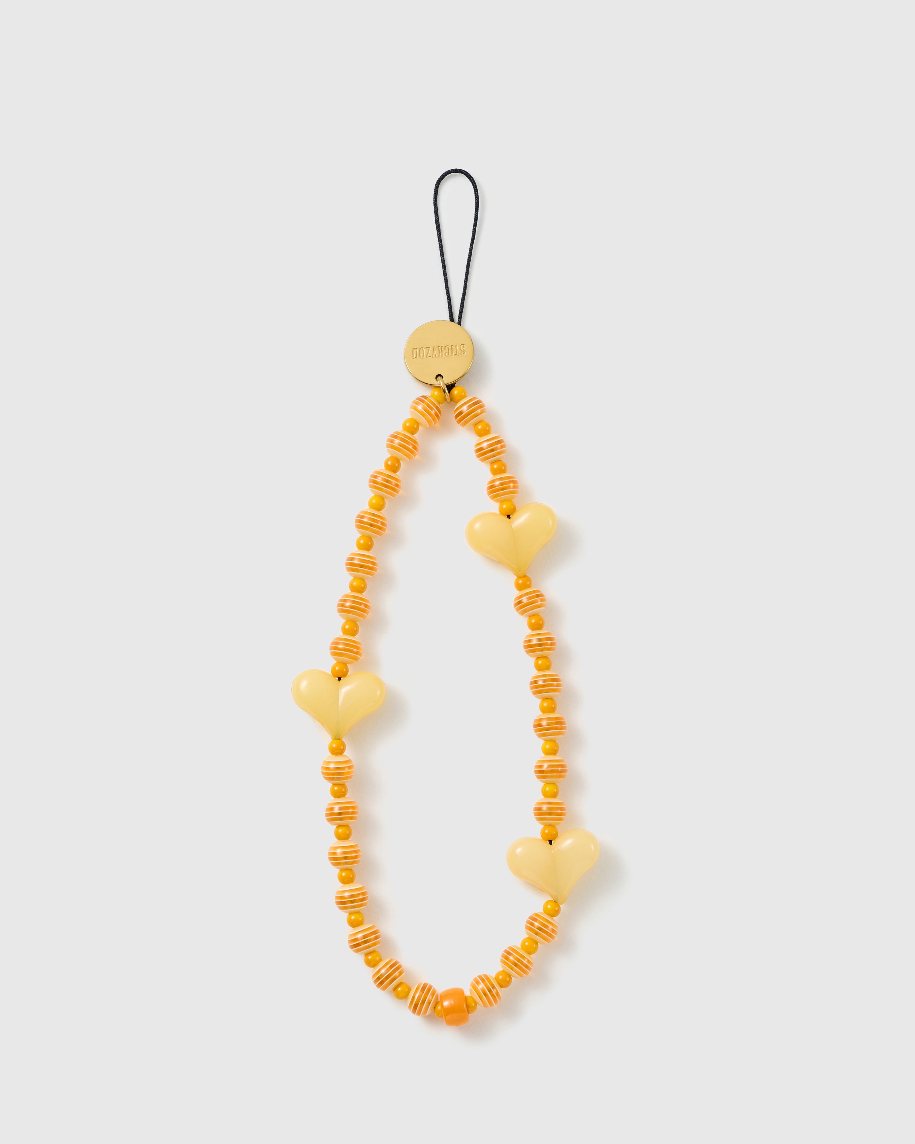STICKYZOO beaded phone strap laid flat: butter-yellow heart beads, orange-and-cream striped beads and a gold-tone engraved disc on a black cord loop.