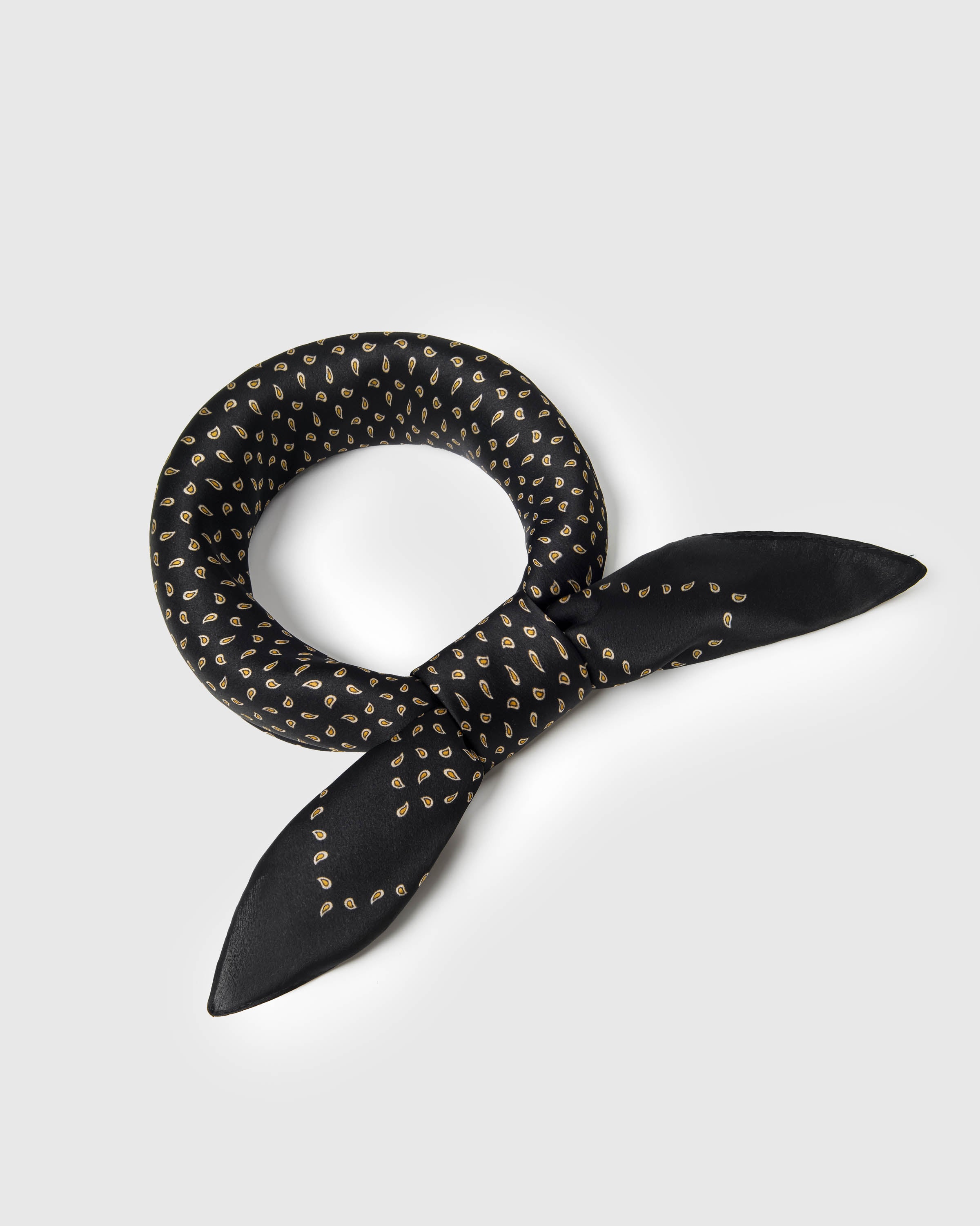 Black silk scarf knotted into a ring with bow tails showing the gold paisley-seed motif on small-square format