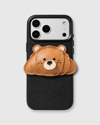 STICKYZOO Croissant Charm Case — handmade 3D leather croissant character charm in warm caramel with layered crescent body, round ears, and black nose on textured black leather phone case. Hand-cut, stitched, and layered from French Alran leather. 5,000+ Reviews.