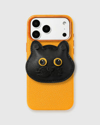Handmade 3D British Shorthair cat charm in black French Alran leather on orange pebbled leather phone case — hand-cut layers, stitched golden eyes, whisker detail, raised camera bezels.