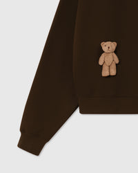 Detail of plush teddy bear near ribbed hem on dark brown Saint Teddy Bear Sweatshirt showing cuff construction