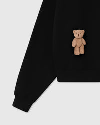 Detail of plush teddy bear near ribbed hem on black Saint Teddy Bear Sweatshirt showing cuff construction
