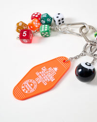 Close-up of translucent red, green, orange, and amber polyhedral dice with white classic dice, black 8-ball charm, and orange Hotel California 1976 motel keychain tag