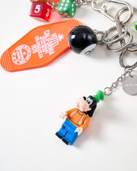 Close-up of collectible mini figure in orange and blue outfit with black 8-ball, orange Hotel California motel tag, colorful dice, and engraved STICKYZOO stainless steel charm tag