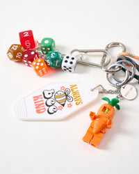 Close-up of orange carrot-suited collectible mini figure, white Always Bee Kind motel tag, colorful polyhedral dice cluster, and STICKYZOO engraved stainless steel tag