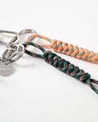 Close-up of dark green and peach hand-braided paracord ropes with intricate knotting pattern, stainless steel carabiner clasp, and engraved STICKYZOO charm tag