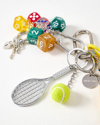 Tennis racket and dice detail | Close-up of silver mini tennis racket, green felt tennis ball, colorful polyhedral gaming dice in yellow, red, green and amber, heart lock with keys, and STICKYZOO engraved stainless steel tag