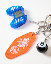 Close-up of translucent blue retro handheld game controller with LCD screen, orange Hotel California 1976 motel keychain tag, and black 8-ball charm on stainless steel chain