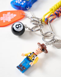 Close-up of collectible cowboy mini figure in yellow shirt and brown hat with black 8-ball charm, STICKYZOO engraved stainless steel tag, and orange motel tag