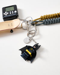 STICKYZOO engraved tag | Close-up of dark caped hero collectible figure with cape and utility belt, STICKYZOO engraved stainless steel charm tag, wood fingerboard deck, and mini game player console
