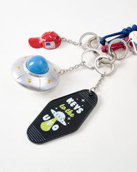 Close-up of black Keys to the UFO motel keychain tag with glow-in-dark UFO illustration, metallic flying saucer with blue dome, and mini red baseball cap charm