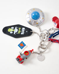 Close-up of metallic UFO saucer with blue dome, rocket pilot figure in red suit holding space flag, Keys to the UFO black motel tag, and engraved STICKYZOO stainless steel charm tag