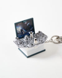 Miniature pop-up book charm opened to reveal sculpted lunar surface with craters, two tiny astronaut figures exploring, and space-themed backdrop illustration