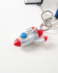 Close-up of retro silver rocket ship charm with red fins, blue cockpit dome, and riveted metallic body connected to stainless steel chain and key rings