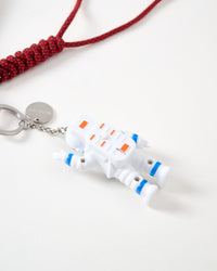 Close-up of white articulated astronaut figure with blue and orange spacesuit details, STICKYZOO engraved stainless steel charm tag, and red hand-braided paracord rope