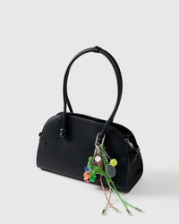 A detail people notice | Dino Stack Charm on black handbag from three-quarter angle showing green dinosaur, rainbow dice, tennis racket and ball, and green and pink braided ropes