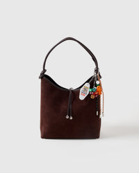 Lucky Harvest Stack Charm clipped onto a dark brown suede hobo bag showing carrot figure, Bee Kind tag, colorful dice, and braided paracord ropes