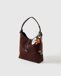 Lucky Harvest Stack Charm on dark brown suede hobo bag from side angle showing carrot figure, Bee Kind tag, dice cluster, and braided ropes with stainless steel tips