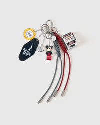 STICKYZOO Lucky Slot Stack Charm flat lay featuring mini slot machine with pull lever, tuxedo character figure, casino 1000-point chip, silver dice, New York City motel tag, and red and black hand-braided paracord ropes with stainless steel metal tips on light gray background