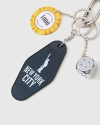Close-up of black New York City motel keychain tag, yellow 1000-point poker chip, and polished chrome dice charm with stainless steel chain links