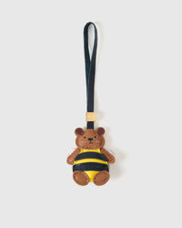 Honeybuzz Bear charm handmade from French Alran genuine goatskin — caramel-brown bear head in a black-and-yellow striped bee costume, translucent wings, tan paws, dark navy leather strap. Three days by hand. Arrives in luxury gift packaging. Designed in Amsterdam.