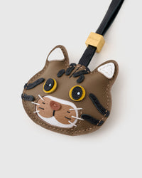 Tight close-up of the brown tabby American Shorthair face — yellow eyes in real leather, bold black stripes hand-stitched across the chestnut forehead, pink nose on caramel muzzle. Every panel by hand.