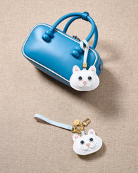 Overhead flatlay — white British Shorthair in strap and keychain editions alongside a blue box bag. Finished one at a time, by hand.