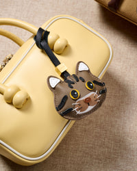 Brown tabby American Shorthair on a pastel-yellow box bag, side angle — 5,000+ reviews across STICKYZOO. Real French leather, 3 days by hand.
