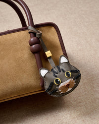 Gray-olive American Shorthair on a tan suede top-handle bag, side angle — real French leather, 3 days by hand.