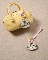 Overhead flatlay — silver-tabby British Shorthair charm with a pastel-yellow box bag and keychain edition. Finished one at a time, by hand.