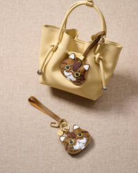Overhead flatlay — Exotic Shorthair in strap and keychain editions alongside a soft-yellow bucket bag. Finished one at a time, by hand.