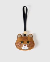 Ginger British Shorthair cat charm handmade from French Alran genuine goatskin — caramel-ginger body, silver-tipped ears, tabby markings, yellow eyes, white chin, black strap. Three days by hand. Arrives in luxury gift packaging. Designed in Amsterdam.