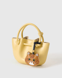 Ginger British Shorthair on a pastel-yellow bucket bag, side angle — real French leather, 3 days by hand.