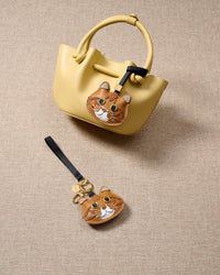Overhead flatlay — ginger British Shorthair charm alongside a pastel-yellow bucket bag and keychain edition. Finished one at a time, by hand.