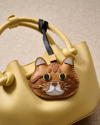 Ginger British Shorthair hanging on a pastel-yellow bucket bag, angled close-up — loved by thousands of cat lovers. Real French leather, 3 days by hand.