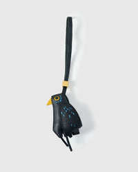Blackbird charm handmade from French Alran genuine goatskin — deep black body, blue hand-stitched feather-dash detail, yellow beak, small yellow-ringed eye, black legs, black leather strap. Three days by hand. Arrives in luxury gift packaging. Designed in Amsterdam.