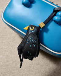 Close-up of the blackbird — blue feather-dash stitches across black goatskin, yellow beak, small ringed eye. Every panel by hand.