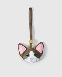 Taupe Devon Rex cat charm — taupe-brown body, white face mask, pink inner ears, green eyes, taupe leather strap. Hand-cut from real French Alran goatskin over three days. Arrives in luxury gift packaging.