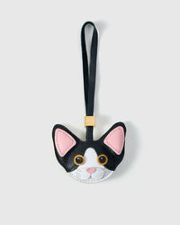 Black-and-white Devon Rex cat charm — black body, white face mask, pink inner ears, gold-orange eyes, black leather strap. Hand-cut from real French Alran goatskin over three days. Arrives in luxury gift packaging.