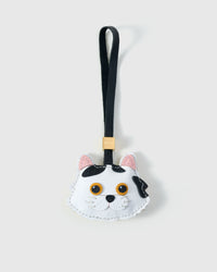 Dragon Li cat charm handmade from French Alran genuine goatskin — white body, bold asymmetric black markings on one side of the face, pink inner ears, gold-orange eyes, black leather strap. Three days by hand. Arrives in luxury gift packaging. Designed in Amsterdam.